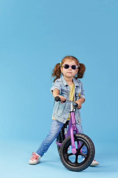 Little girl learns to keep balance while riding a bicycle. Stylish child in sunglasses  isolated on blue background - Foto, imagen