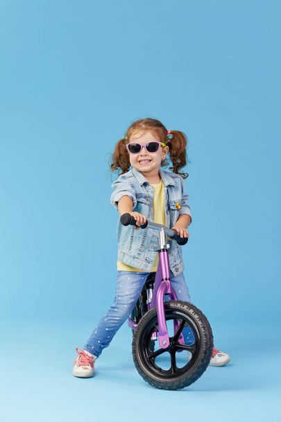 Ein kleines Mädchen lernt beim Fahrradfahren das Gleichgewicht zu halten. stylisches Kind mit Sonnenbrille auf blauem Hintergrund - Foto, Bild