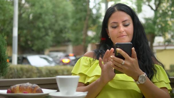 Beautiful brunette woman using cellphone at coffee shop outdoors. Breakfast with a croissant and coffee, checking the news on your smartphone. - Foto, imagen