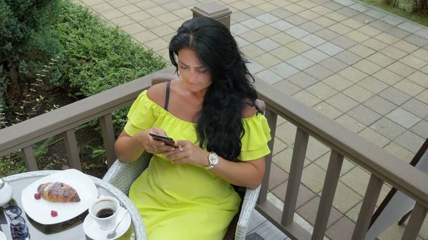 Beautiful brunette woman using cellphone at coffee shop outdoors. Breakfast with a croissant and coffee, checking the news on your smartphone. - Фото, зображення