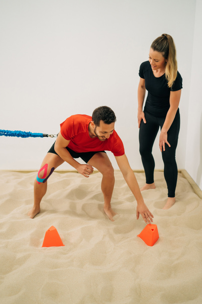 Een jongen met een blauwe steun aan de taille doet stretching oefeningen op zand, bijgestaan door een fysiotherapeutisch meisje, een witte muur op de achtergrond. Concept van spiergezondheid en spierontspanning. - Foto, afbeelding