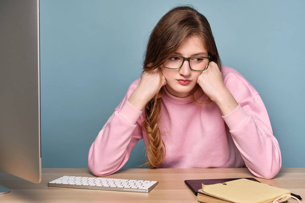 A girl in a pink sweater sits at a table with a computer and books, propping her head with her fists looking to the side. - Photo, image