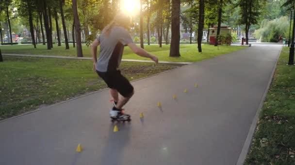 Young long-haired bearded man roller skater is dancing between cones in a nice evening in a city park. Freestyle slalom Roller skating between cones in slow motion. - Séquence, vidéo