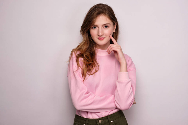 A girl in a pink sweater stands on a white background, looks at the camera, propping her cheek with her finger. - Foto, Imagen