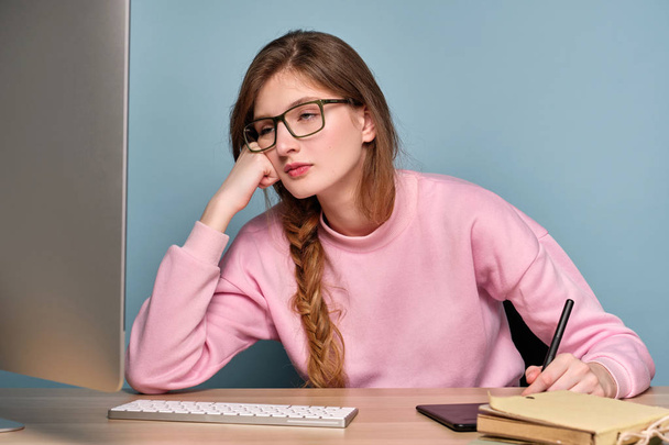 A girl in a pink sweater and glasses sits at a computer and looks in surprise at the monitor with a paper cup in her hand. - Fotografie, Obrázek