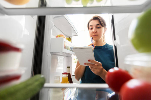 woman making list of necessary food at home fridge - Foto, Imagen