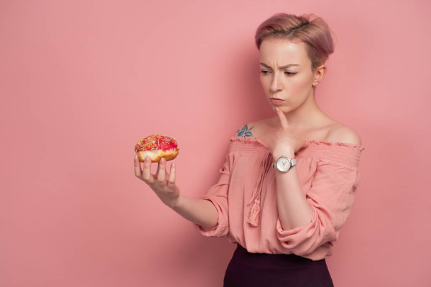 A girl with short pink hair, holds a donut in her hand, and frowns at him, on a pink background. - Фото, изображение