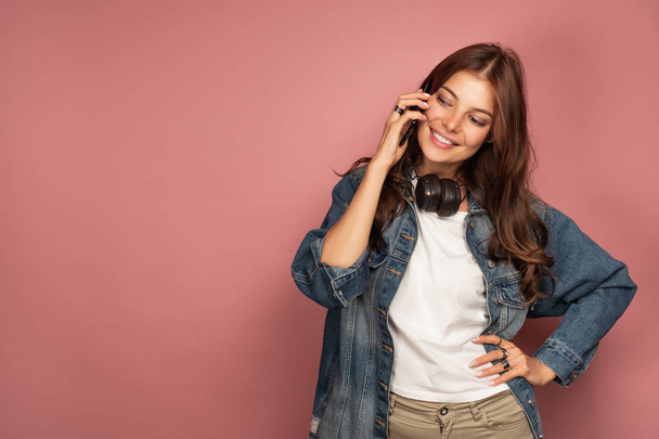 A girl in jeans stands on a pink background with headphones on her neck and speaks on the phone with a smile looking down - Фото, изображение