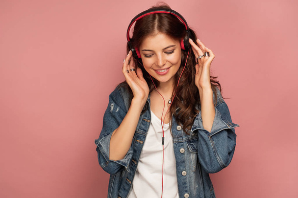 A dark-haired girl in a jeans shirt stands on a pink background clutching headphones and smiles with her head down. - Φωτογραφία, εικόνα