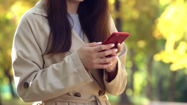 Młoda kobieta ubrana w płaszcz lub rów za pomocą smartfona stojącego w jesiennym parku - Materiał filmowy, wideo