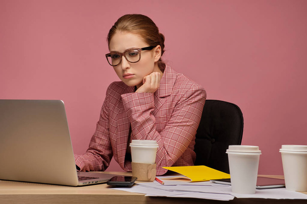 Redhead girl in a business suit and glasses on a pink background sitting at a table and looking thoughtfully at a laptop. - 写真・画像