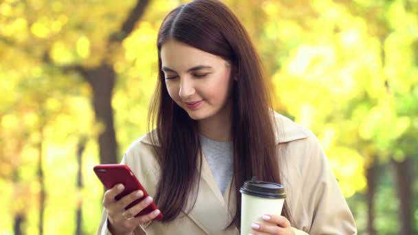 Mujer al aire libre usando un abrigo o trinchera en el parque con su café y teléfono celular
 - Imágenes, Vídeo