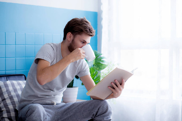 Νέος άνθρωπος διαβάζοντας βιβλίο και πίνοντας καφέ στον καναπέ στο ζωντανό - Φωτογραφία, εικόνα