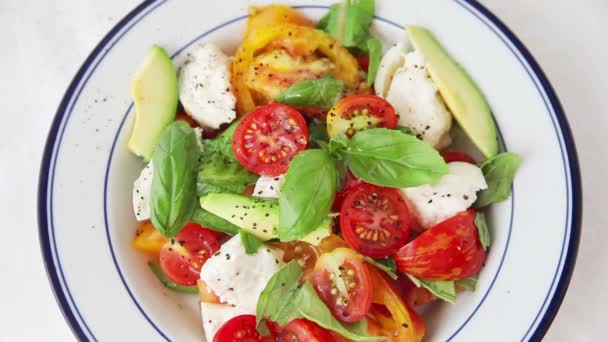 Olijfolie en een vork worden toegevoegd aan een verfrissende zomer salade van avocado, tomaten, zachte kaas, verse basilicum en zwarte peper - Video