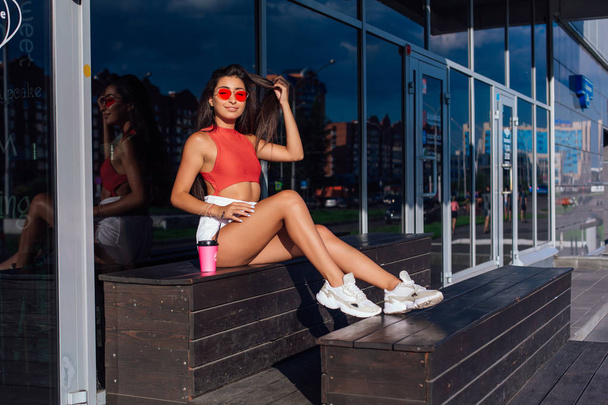 Stylish happy young brunette woman wearing white shorts and sneakers holding pink cup of coffee to go sitting next to coffee shop. - Foto, Imagem