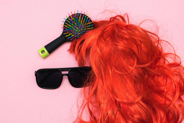 Черные очки, расческа и оранжевый парик на розовом фоне. Аксессуары для создания стиля
. - Фото, изображение