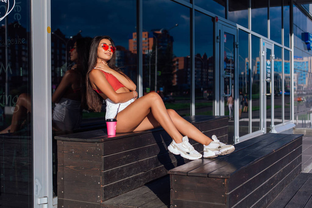Stylish happy young brunette woman wearing white shorts and sneakers holding pink cup of coffee to go sitting next to coffee shop. - Fotó, kép