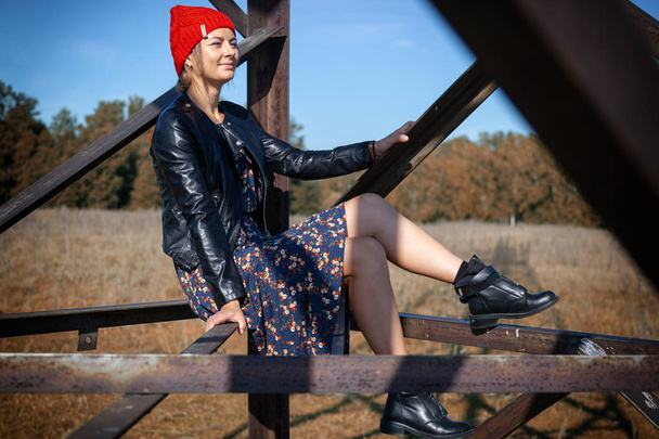 Portrait of a  cheerful woman in a knitted hat,  leather coat, pretty dress and boots, smiles cute, poses, smiles and sits on birch logs. The concept of rustic style and life in ancient times - Photo, Image