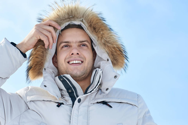 Fashion young beautiful man in a white jacket with fur hood in winter - Фото, зображення