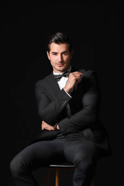 Fashionable man in formal clothes sitting on chair against dark background - Фото, зображення