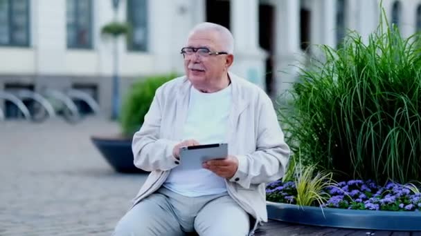 senior man with tablet sits at flowerbed on city square - Video
