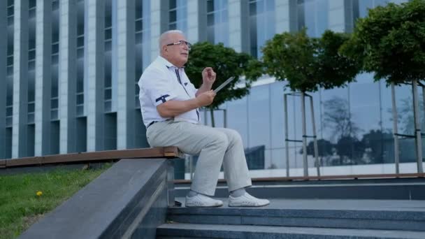 cheerful pensioner holds tablet sitting on street bench - Metraje, vídeo