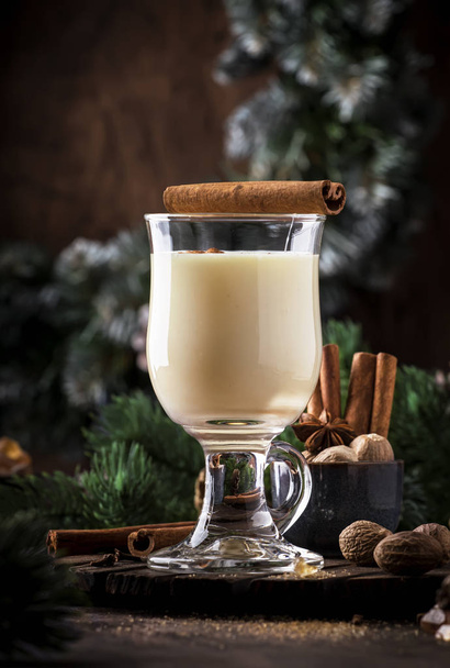 New Year or Christmas Eggnog cocktail - hot winter or autumn drink with milk, eggs and dark rum, sprinkled with cinnamon and nutmeg in a glass on wooden background, festive decoration - Photo, Image