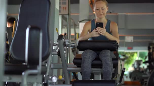 A young woman at the gym doing exercises holding a phone in front of her eyes in her hands. All the attention is in the phone. The concept of dependence on social networks. Mobile addiction concept - Кадры, видео