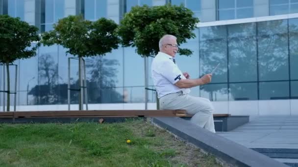 senior man uses tablet on bench at modern office center - Кадры, видео