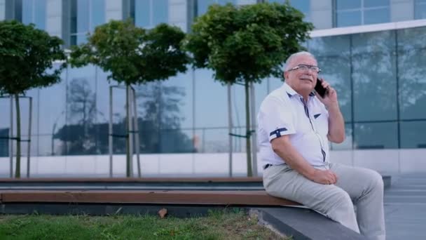 senior businessman talks on phone resting on bench in city - Video