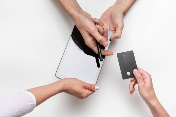 Close up top view of a woman hands over bank card to a man in order to pay for the product. Tablet and pen on a white table. Concept of e-commerce and online shopping. Successful deal. - 写真・画像