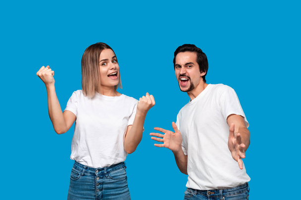 Happy couple expressing emotion of success. Young blond woman and bearded man with mustaches in white tees and blue jeans are winner. Models standing isolated over blue background. - Photo, Image