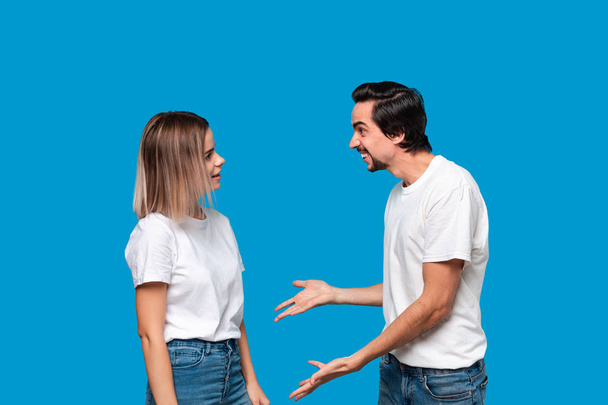 Paar einer jungen blonden Frau und eines brünetten bärtigen Mannes mit Schnurrbart in weißen T-Shirts und blauen Jeans, die sich vor blauem Hintergrund streiten. - Foto, Bild