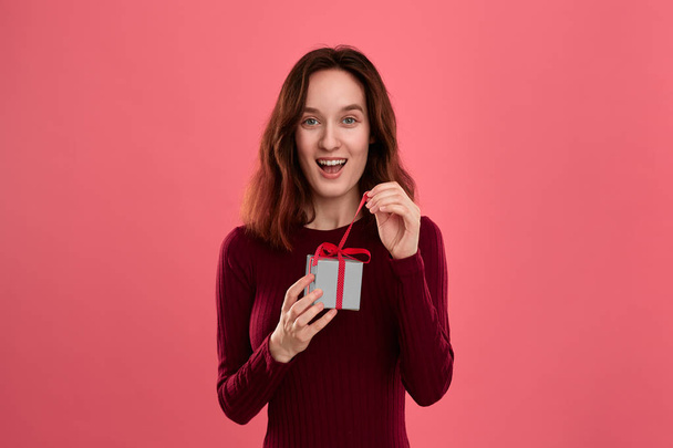 Excited pretty brunette girl opens gift packed in present box with a ribbon standing isolated on a dark pink background and smiling at the camera. Celebrating special event. - Photo, Image
