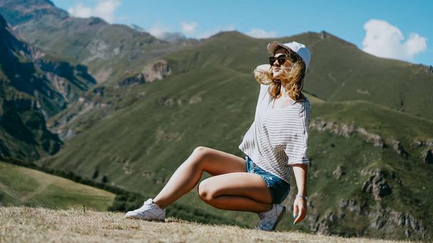 The girl in a cap and shorts in the mountains sits on the grass - Foto, Imagem