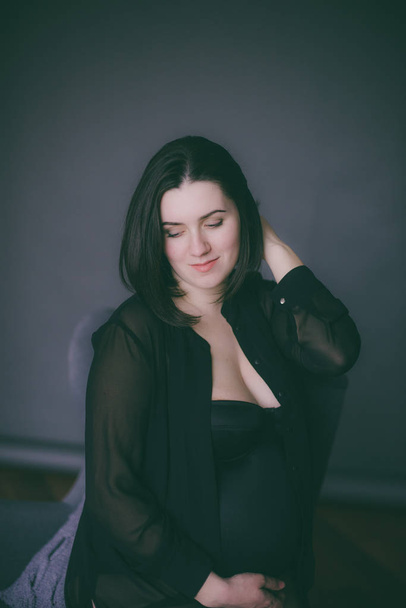 Young pregnant woman with dark hair in black lingerie and a black silk shirt on a dark background - Photo, Image