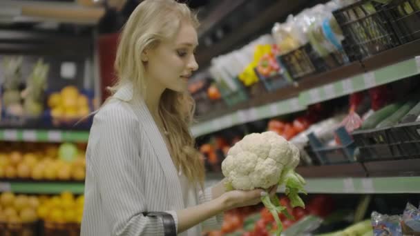 Молодая красивая брюнетка в возрасте 20 лет выбирает капусту и цветную капусту и кладет их в корзину для покупок в проходе с фруктами и овощами в продуктовом магазине
. - Кадры, видео