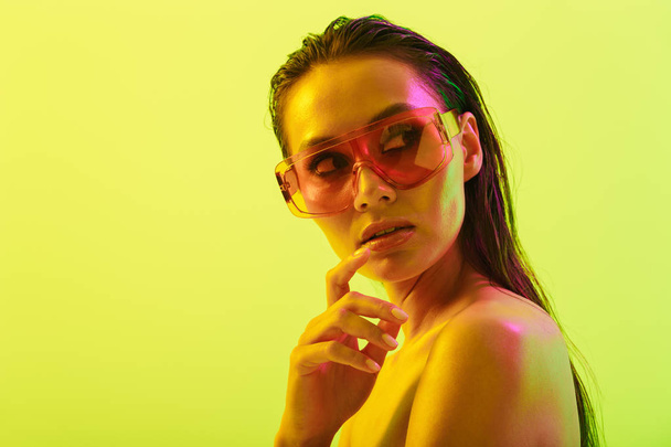 Amazing young woman posing isolated over yellow wall background with neon bright lights wearing fashion sunglasses. - Foto, imagen