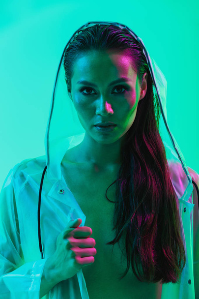 Serious woman posing isolated over blue wall background with neon bright lights dressed in raincoat. - 写真・画像