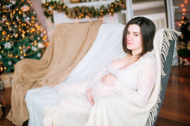 Young pregnant girl with dark hair in a lace white dress on the sofa in a Christmas setting - 写真・画像
