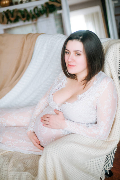 Young pregnant girl with dark hair in a lace white dress on the sofa in a Christmas setting - Photo, image