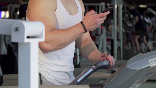 Hombre irreconocible usando un teléfono inteligente, mientras camina en una cinta de correr en el gimnasio
 - Foto, Imagen