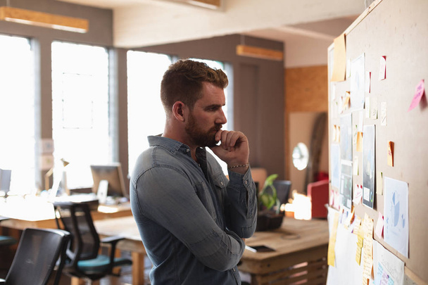 Vista lateral de perto de um jovem caucasiano lendo notas em uma parede e pensando durante uma sessão de brainstorm em equipe em um escritório criativo
 - Foto, Imagem