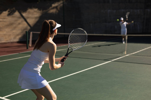 Rear view of a young Caucasian woman and a man playing tennis on a sunny day, man hitting a ball - Photo, Image