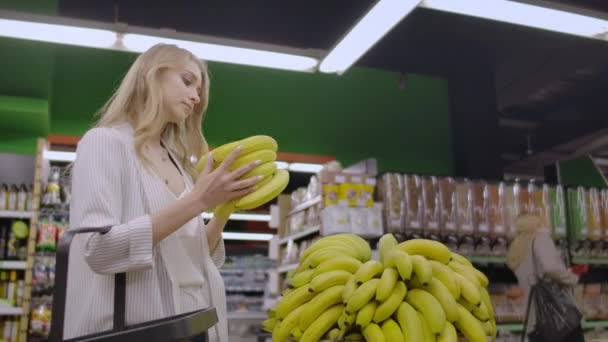 Mladá krásná brunetka ve dvaceti si vybírá banány a oblékala je do nákupního vozíku u ovocné a zeleninové uličky v potravinách. - Záběry, video