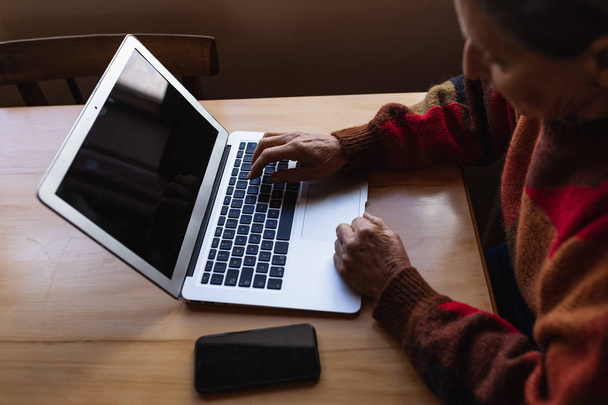 Yan görünüm, yanında akıllı telefon olan bir dizüstü bilgisayar kullanarak evde ki bir masada oturan üst düzey bir Beyaz kadının yan görünümü - Fotoğraf, Görsel