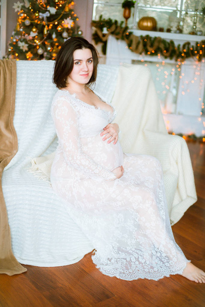 Young pregnant girl with dark hair in a lace white dress on the sofa in a Christmas setting - Fotoğraf, Görsel