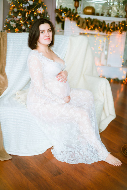 Young pregnant girl with dark hair in a lace white dress on the sofa in a Christmas setting - Photo, image