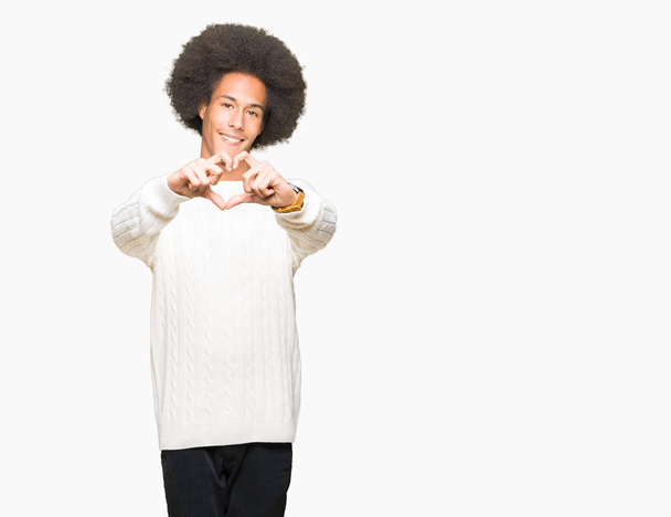 冬のセーターを着たアフロヘアの若いアフリカ系アメリカ人男性が、ハートのシンボルと形を手で見せる恋に微笑む。ロマンチックなコンセプト. - 写真・画像