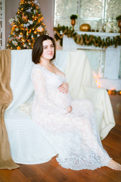 Young pregnant girl with dark hair in a lace white dress on the sofa in a Christmas setting - Foto, Imagem